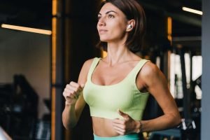 2127462771-fit-caucasian-beautiful-woman-fitting-sport-wear-gym-running-jogging-treadmill-with-wireless-headphones-ears-scaled-1